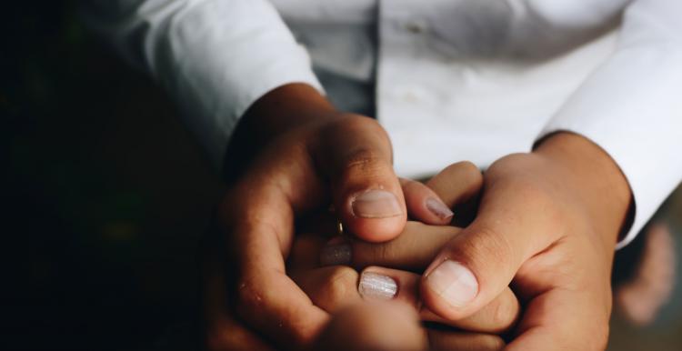 One person is holding their hand together and another person has placed their hand around them to show support.  