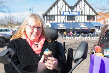 Woman in an electric wheelchair is holding a cupcake while outside.