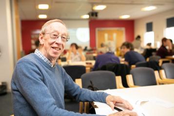 An older man sitting joyfully at a table in a communal space with other elderly patients. 