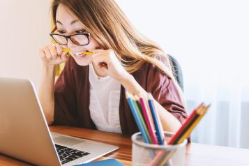 A woman is biting a pencil while looking at the content on her laptop