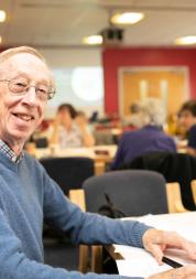 An older man sitting joyfully at a table in a communal space with other elderly patients. 